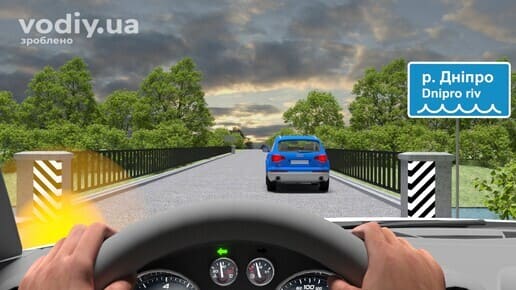 Синій автомобіль на мосту, дорожній знак 5.65.1 «Назва об’єкта» р. Дніпро, увімкнений лівий покажчик повороту на панелі приладів водія.
