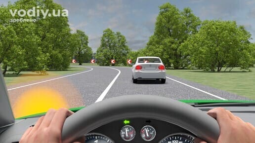 Білий автомобіль на дорозі з крутим поворотом, дорожні знаки 1.4.1 «Напрямок повороту», обмежена оглядовість через дерева.