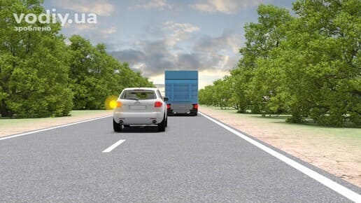 Білий легковий автомобіль з увімкненим лівим покажчиком повороту готується до обгону вантажівки на дорозі поза населеним пунктом.