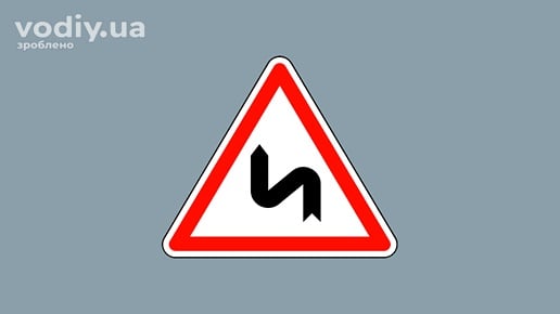 Дорожній знак 1.3.2 «Декілька поворотів» з першим поворотом ліворуч на сірому фоні.