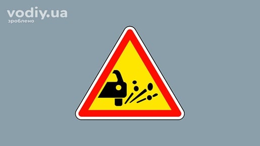 Дорожній знак 1.14 «Викидання кам’яних матеріалів» на жовтому фоні, попередження про гравій та щебінь на дорозі.