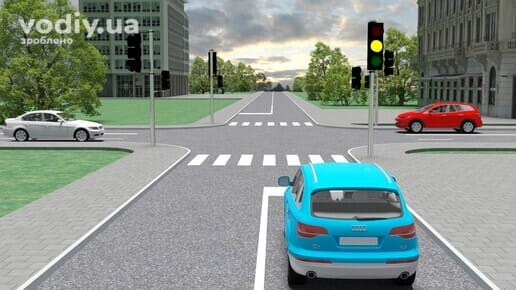 Блакитний автомобіль перед перехрестям, жовтий сигнал світлофора, пішохідний перехід та край проїзної частини.