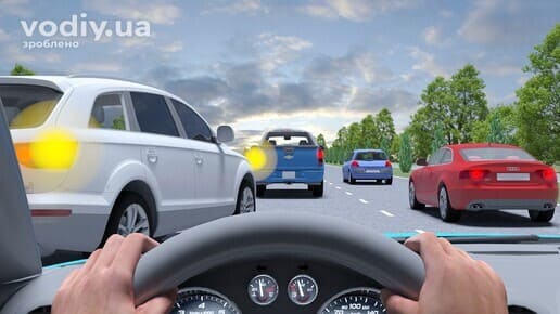 Білий автомобіль з увімкненим правим покажчиком повороту перестроюється перед вашим авто, рух по багатосмуговій дорозі поза населеним пунктом.