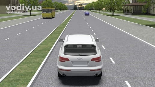 Білий автомобіль на дорозі з розділювальною смугою, автобус та синє авто попереду, рух по крайній лівій смузі поза населеним пунктом.