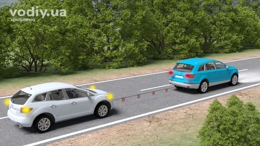 Буксирування синім автомобілем білого кросовера на гнучкому зчепленні за межами населеного пункту, увімкнена аварійна сигналізація.