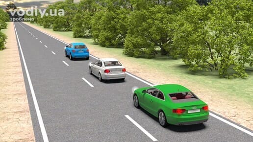 Зелений автомобіль з увімкненим лівим покажчиком повороту готується до обгону білого та синього авто на заміській дорозі.